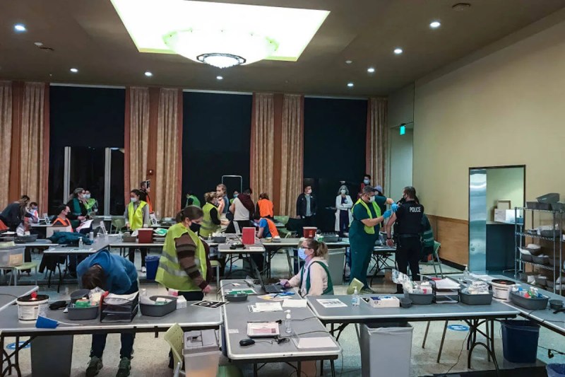 Healthcare workers in protective equipment administer vaccines to patients in a conference room with folding tables. It is after sunset.