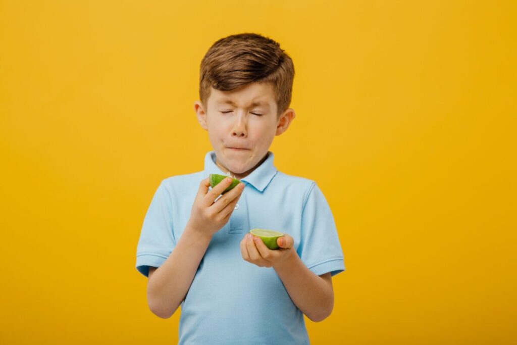 Little boy taste lime, sour taste, makes grimace, facial emotions negative, in blue T-shirt, isolated yellow background, copy space.