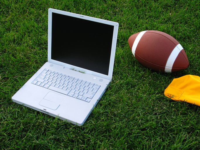 This color photo shows a closeup view of a white laptop computer, a football, and a bright-yellow penalty flag on a deep-green grassy football field in an attempt to create a fantasy football concept image. The grassy turf fills the entire background of the image.