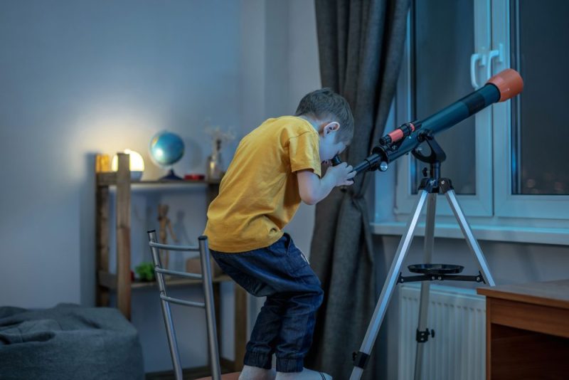 Young boy is looking through a telescope in a room at the night starry sky.